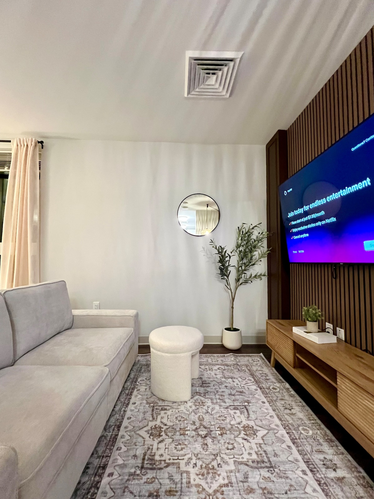 A modern living area features a light gray sofa positioned near a wooden media console. A round white ottoman complements the space. A circular mirror is mounted on the wall, reflecting light, while a decorative plant adds a touch of greenery. A smart TV mounted nearby displays content options.
