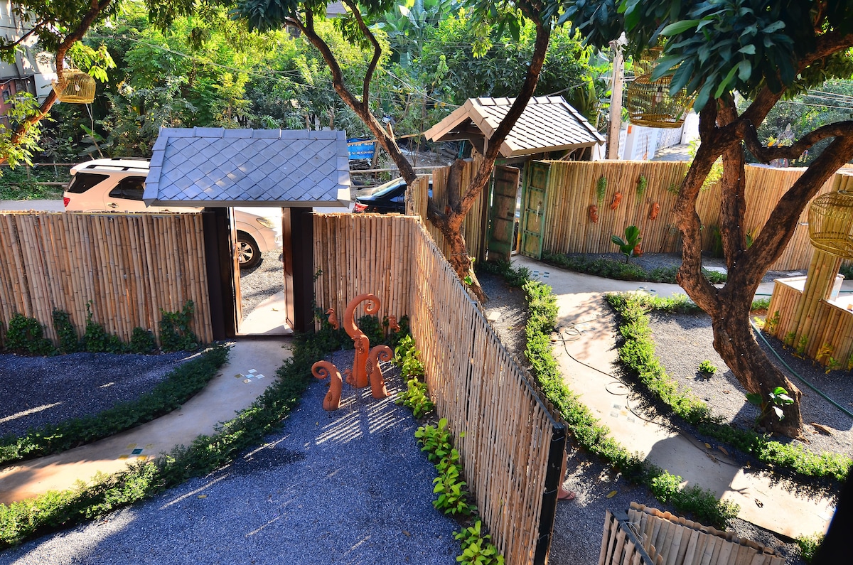 A tranquil garden space is enclosed by bamboo fencing, featuring winding pathways lined with lush greenery. A decorative orange sculpture is visible along the path, with a wooden gate leading to the entrance. The area offers a serene atmosphere amid surrounding foliage.
