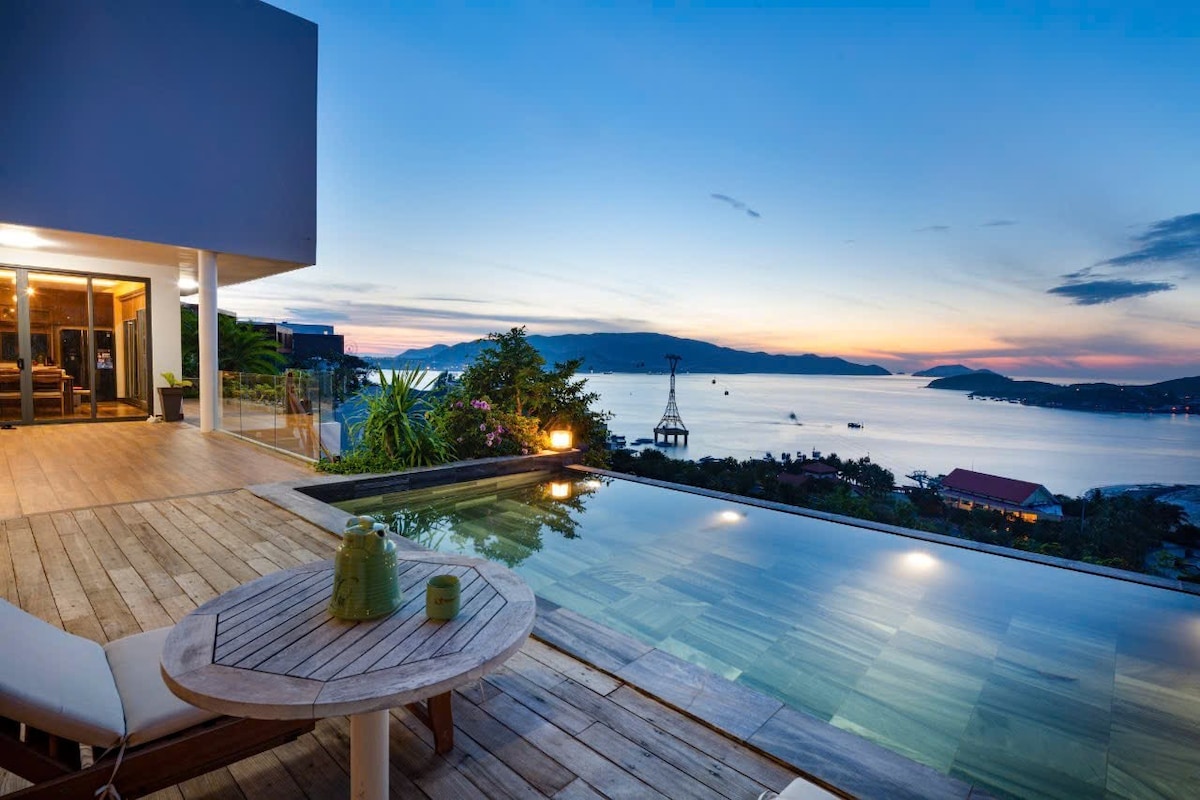 An outdoor terrace showcases a serene private pool reflecting the evening sky, surrounded by lush greenery. A wooden table is set with two green glasses, inviting relaxation. The expansive view of the bay and distant hills contributes to a calming atmosphere.