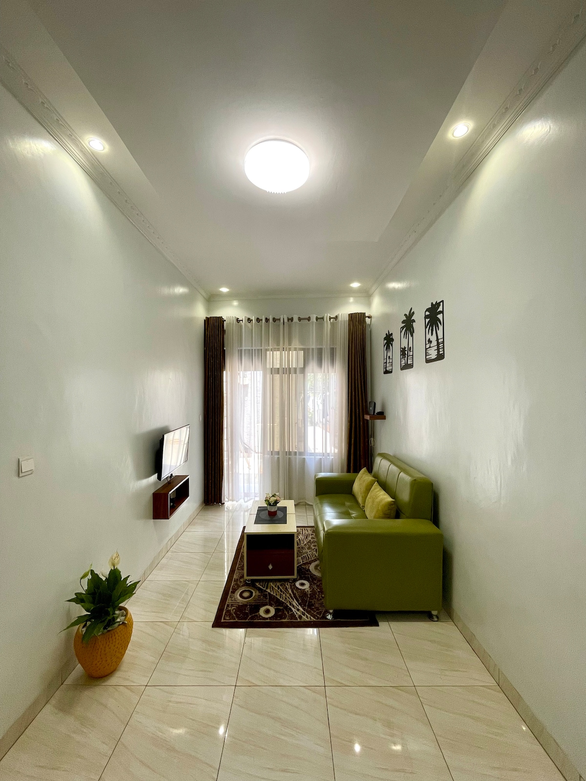 A well-lit living room features a modern L-shaped green sofa facing a sleek TV unit. A stylish center table is placed on a patterned rug. Natural light flows in from large windows, adorned with sheer curtains, enhancing the airy feel of the space.