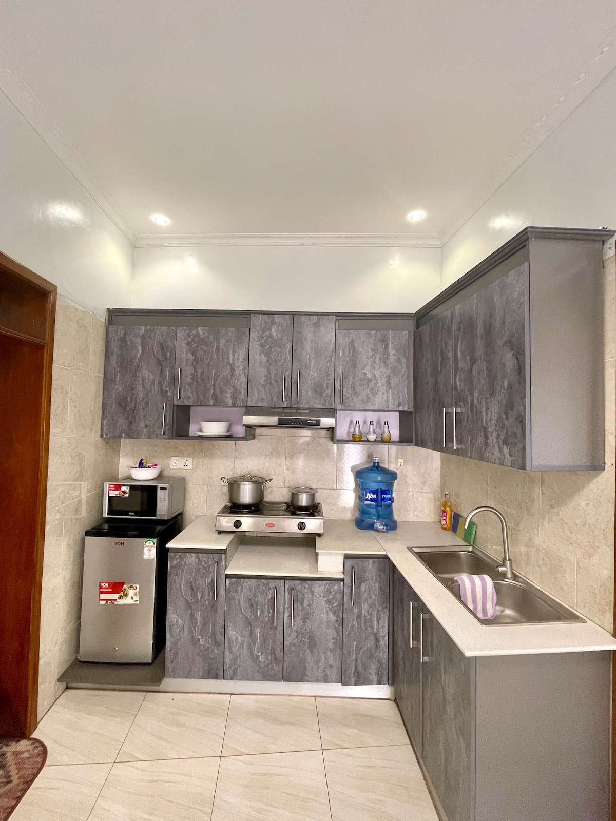 A modern kitchen area is shown, featuring gray cabinetry and sleek countertops. Essential appliances include a refrigerator and a stove with two burners. A blue water dispenser sits prominently, with various kitchen utensils and a sink visible, ensuring a functional cooking environment.