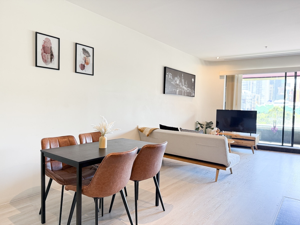 A modern dining area features a dark table surrounded by four brown chairs. A comfortable beige sofa sits nearby, facing a television against a light wall, while two framed art pieces add a touch of decor. Large windows allow natural light to enhance the open space.