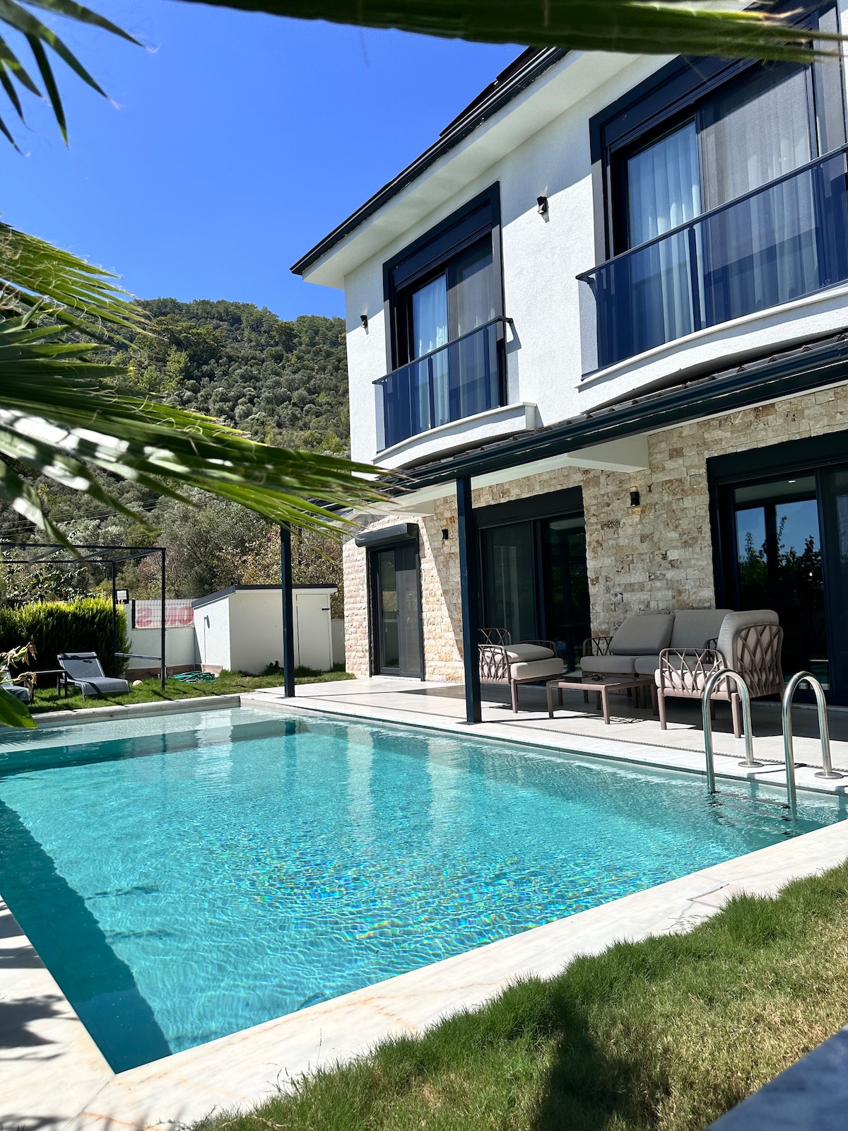 A private pool is framed by a stone patio and manicured grass, offering a serene outdoor space. The building showcases large windows and balconies, allowing ample natural light. Comfortable seating arrangements are arranged under the overhang, providing a relaxing spot to enjoy the view.