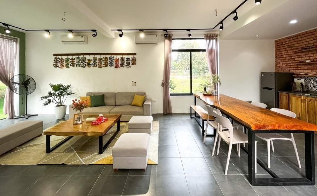 A spacious living area features a light grey sectional sofa and a wooden dining table with white chairs. Natural light floods the room through large windows, highlighting the wooden accents in the kitchen area and a decorative wall with hanging art.