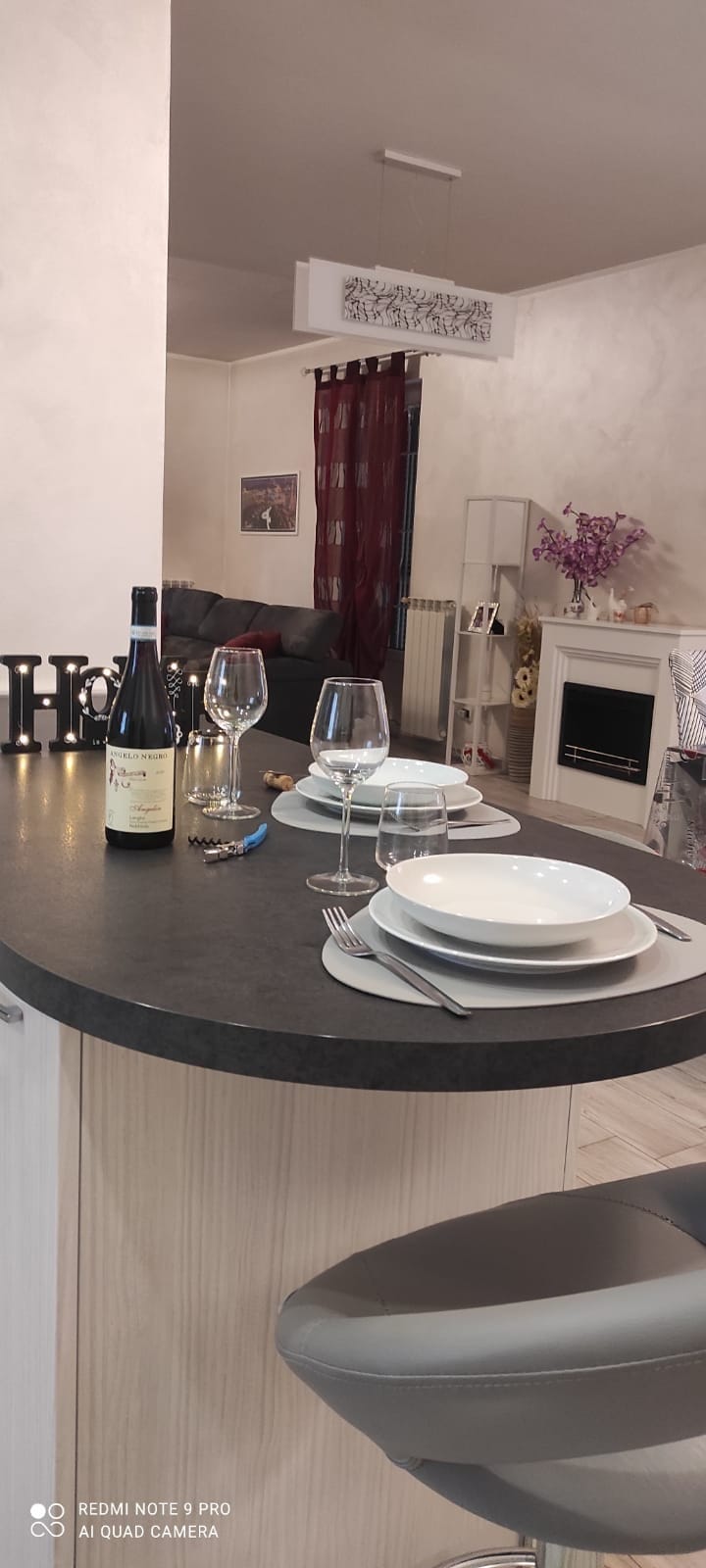 A modern kitchen counter is set for dining, featuring elegant tableware including plates and wine glasses. A bottle of wine stands in the center, with subtle lighting creating a warm ambience. In the background, a living area with a cozy sofa and decorative elements is visible.