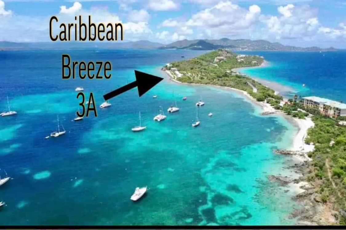 Aerial view of a vibrant Caribbean coastline features clear turquoise waters with multiple sailboats dotting the sea. Lush green land extends towards the horizon, with a distant view of a scenic island. The text indicates the location as 'Caribbean Breeze 3A.'