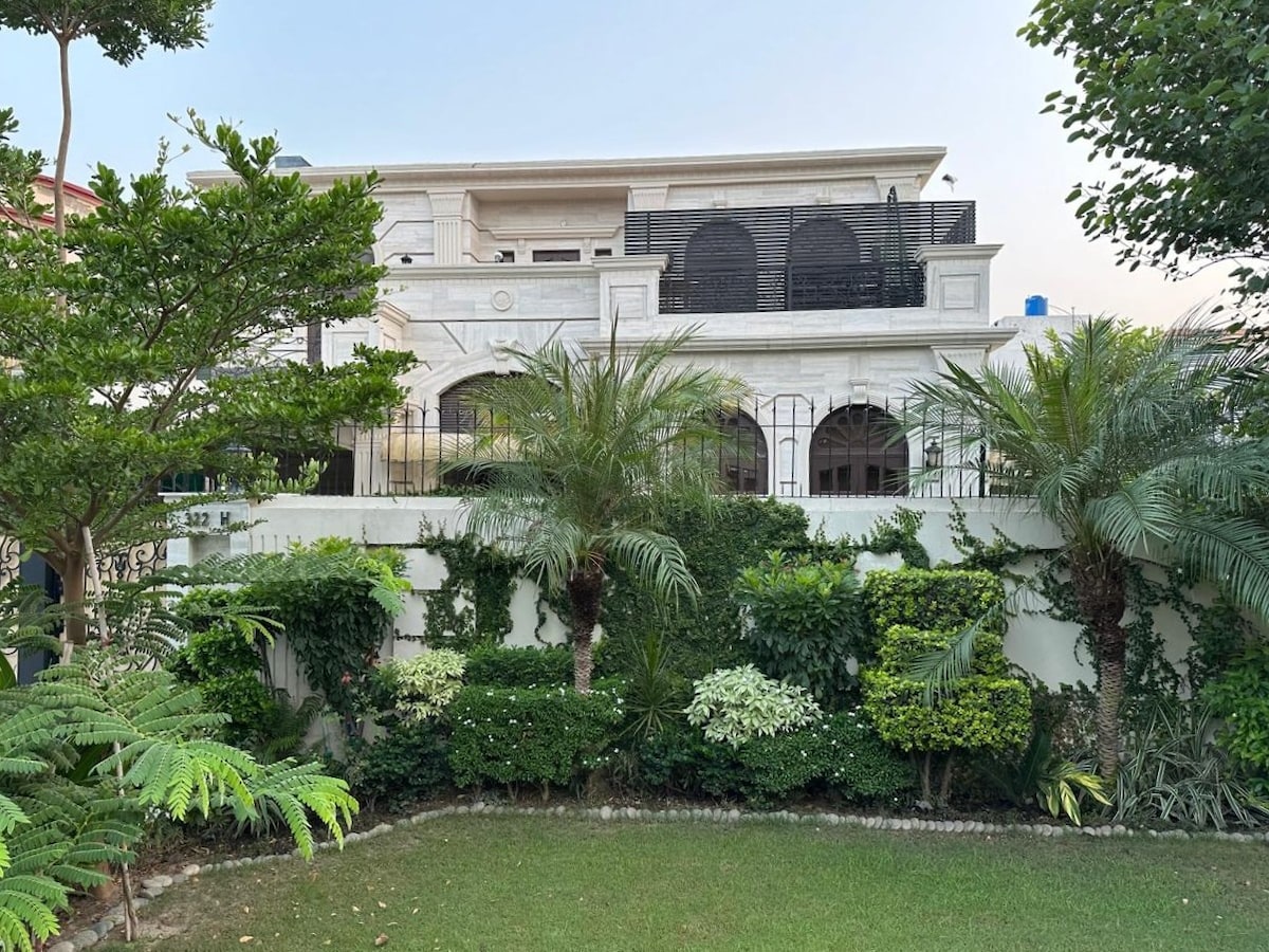 A serene outdoor view highlights a spacious property surrounded by well-maintained greenery. Lush plants and manicured hedges frame the structure, which features arched balconies and large windows, inviting natural light into the upper levels.