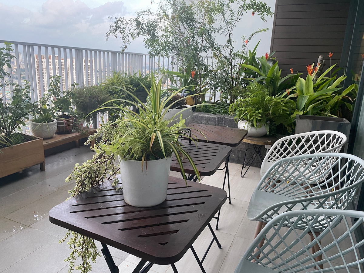 A spacious balcony is adorned with lush greenery and various potted plants. Two tables made of dark wood are positioned alongside stylish light blue chairs, creating an inviting outdoor seating area. The balustrade offers a glimpse of the cityscape in the background.