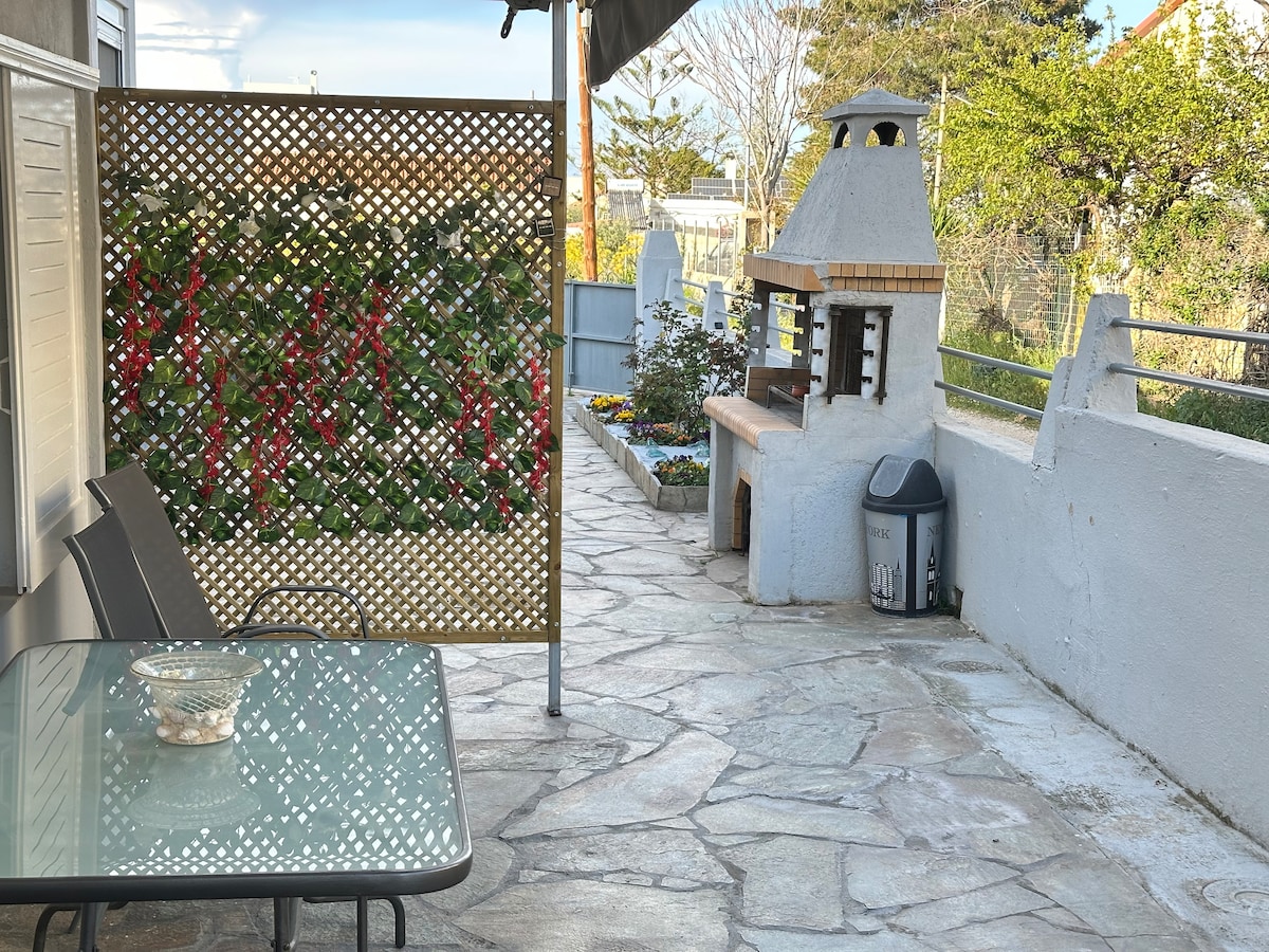 An outdoor patio area is designed with a stone floor, featuring a glass table alongside two chairs. A built-in barbecue grill is situated against a wall, with a small garden area surrounded by green foliage and colorful flowers in the background.