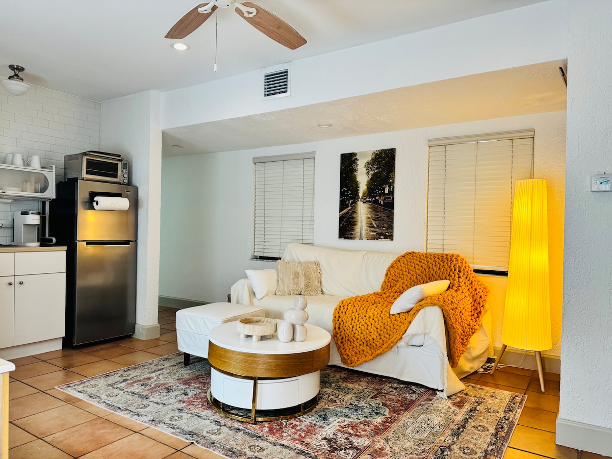 A bright living area features a white sofa adorned with a textured orange throw. A circular coffee table is centered on a patterned area rug, with decorative items placed on top. Natural light filters through the window, complemented by stylish furnishings and a standing lamp.