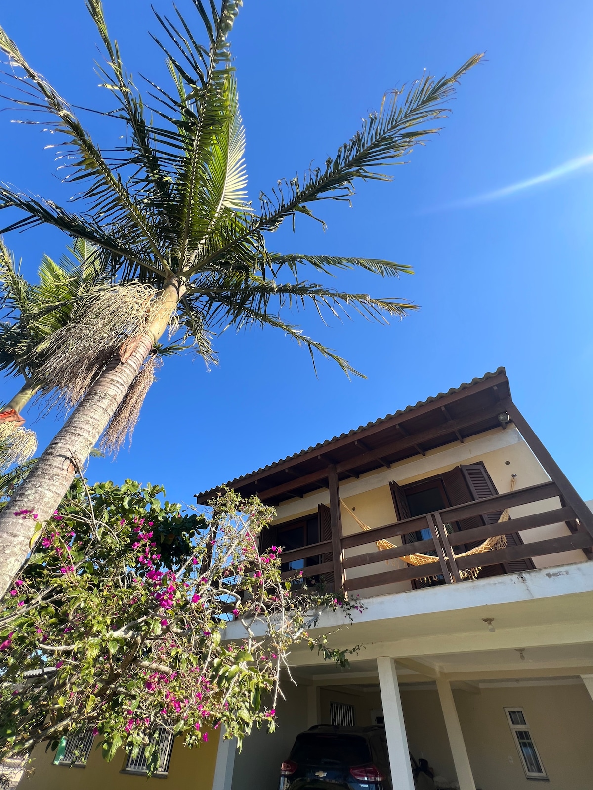 A two-story house is shown with a wooden balcony and brown shutters. A palm tree with green fronds is visible beside the house, complemented by vibrant pink flowers. The clear blue sky creates a bright atmosphere, while a vehicle is parked beneath the home.