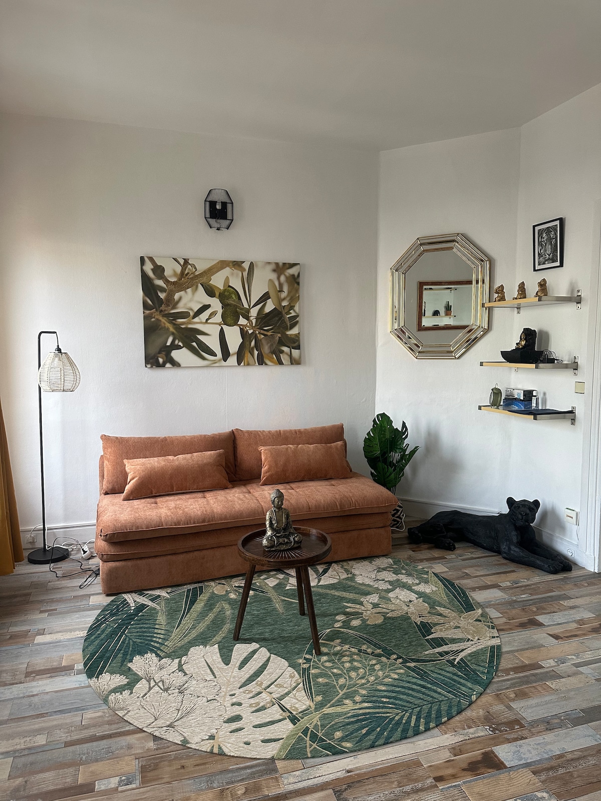 A cozy living area is depicted, showcasing an orange sofa contrasted by a green patterned rug. A decorative mirror and shelves with various items are mounted on the wall. A small table with a statue sits centrally, while a dark figure resembling a panther rests beside a decorative plant.