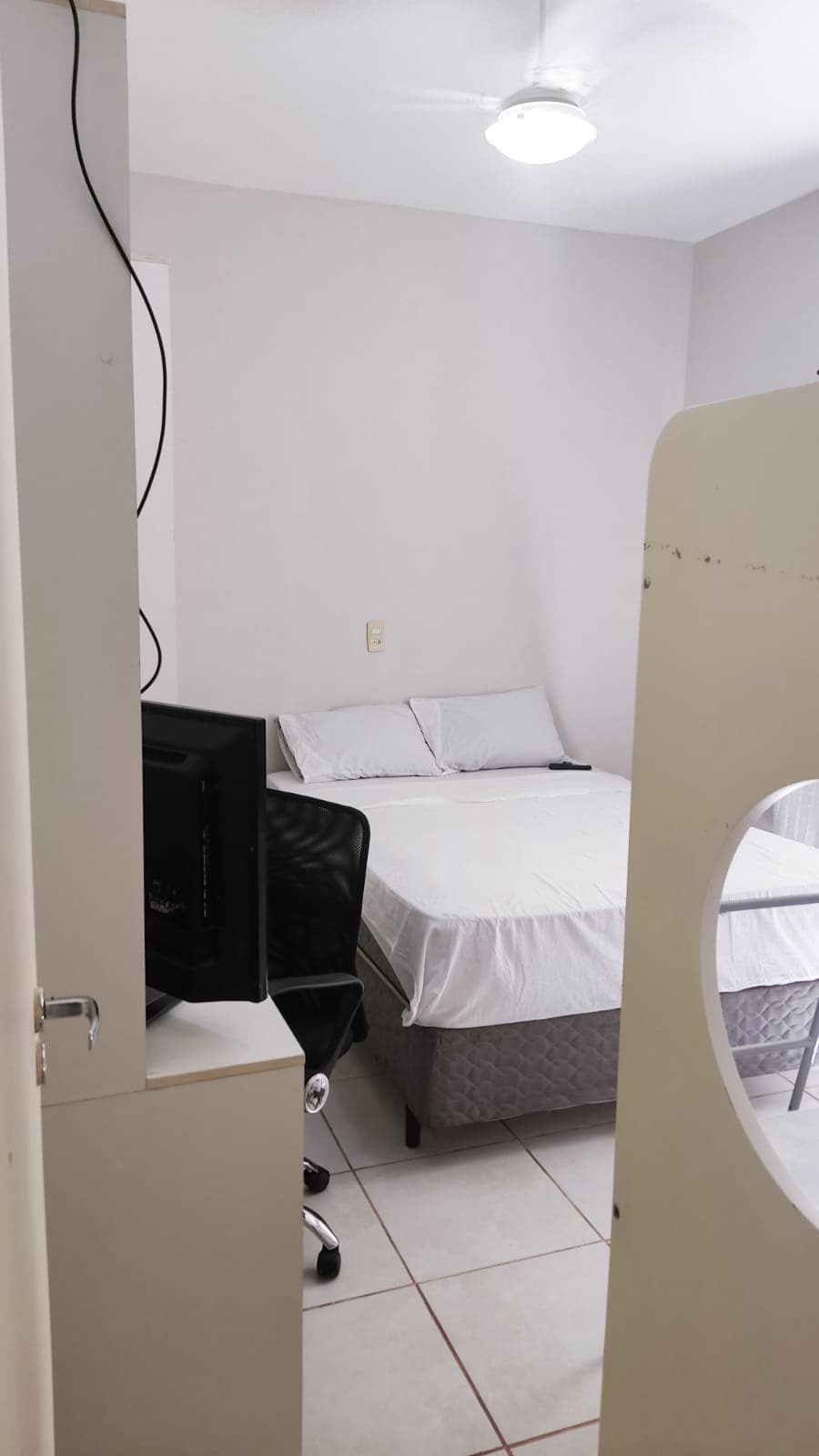 A cozy bedroom is visible, featuring a spacious bed with light bedding and a small desk with a black chair. A television is positioned on a nearby cabinet. The room has neutral-colored walls and a ceiling fan, bathed in natural light from a partially obscured window.