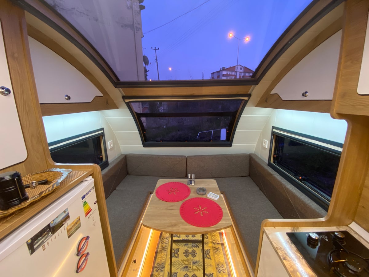 The interior of the caravan is displayed, featuring a spacious seating area with a grey couch and a wooden table at the center. Large windows allow natural light, and modern appliances are visible, including a small kitchenette with a sink. The warm lighting creates a cozy environment.