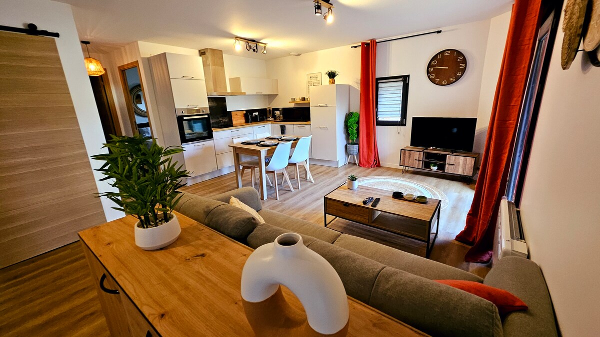 A cozy living space features a grey sofa facing a wooden coffee table. An open kitchen with modern appliances and a dining table for four is visible. Natural light floods the area through large windows, complemented by vibrant red curtains.