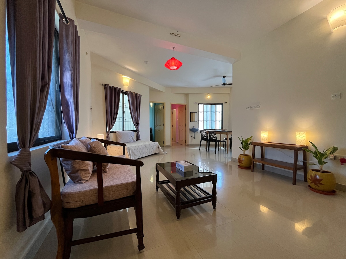 A spacious living area features large windows allowing natural light to fill the room. Comfortable seating is provided by a wooden sofa and chairs, while a coffee table anchors the space. The walls are dressed in soft tones, complementing the warm lighting fixtures.