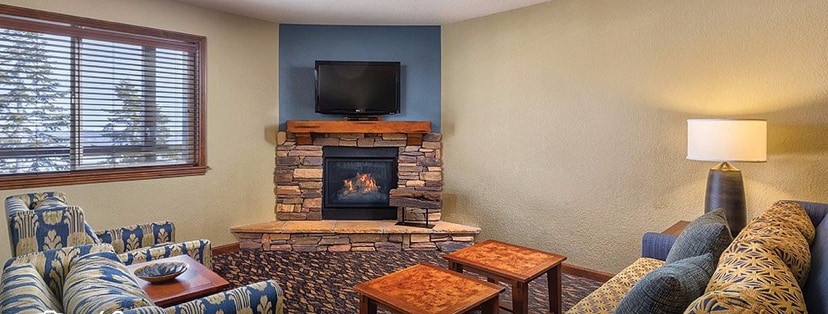 A welcoming living area features a cozy stone fireplace and a flat-screen TV mounted on the wall. Comfortable seating is provided by patterned chairs and a spacious sofa. Natural light filters through window coverings, illuminating the rich textures of the carpet and wooden tables.