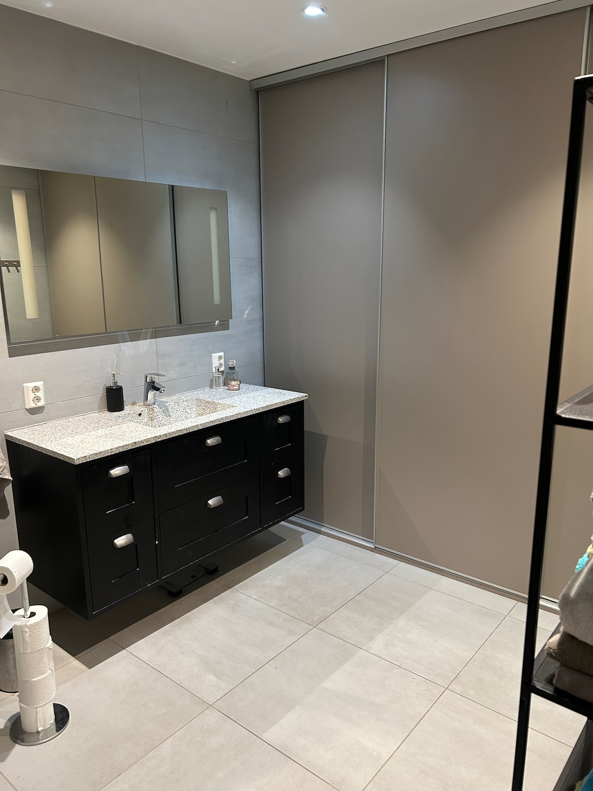 A modern bathroom features a dark cabinet with multiple drawers beneath a gray granite countertop. A large mirror is mounted above the sink, reflecting the sleek design. Neutral-toned tiles cover the floor, and a grey sliding door is positioned on one side.
