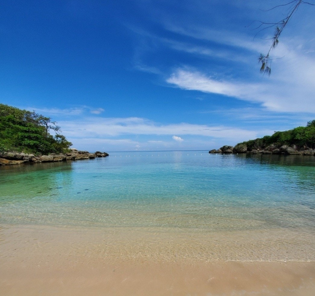 A calm beach scene features clear, shallow waters gently lapping at a sandy shore. The horizon blends a bright blue sky with soft clouds, while lush greenery lines the edges of the cove. Rocky formations provide natural framing to the tranquil landscape.