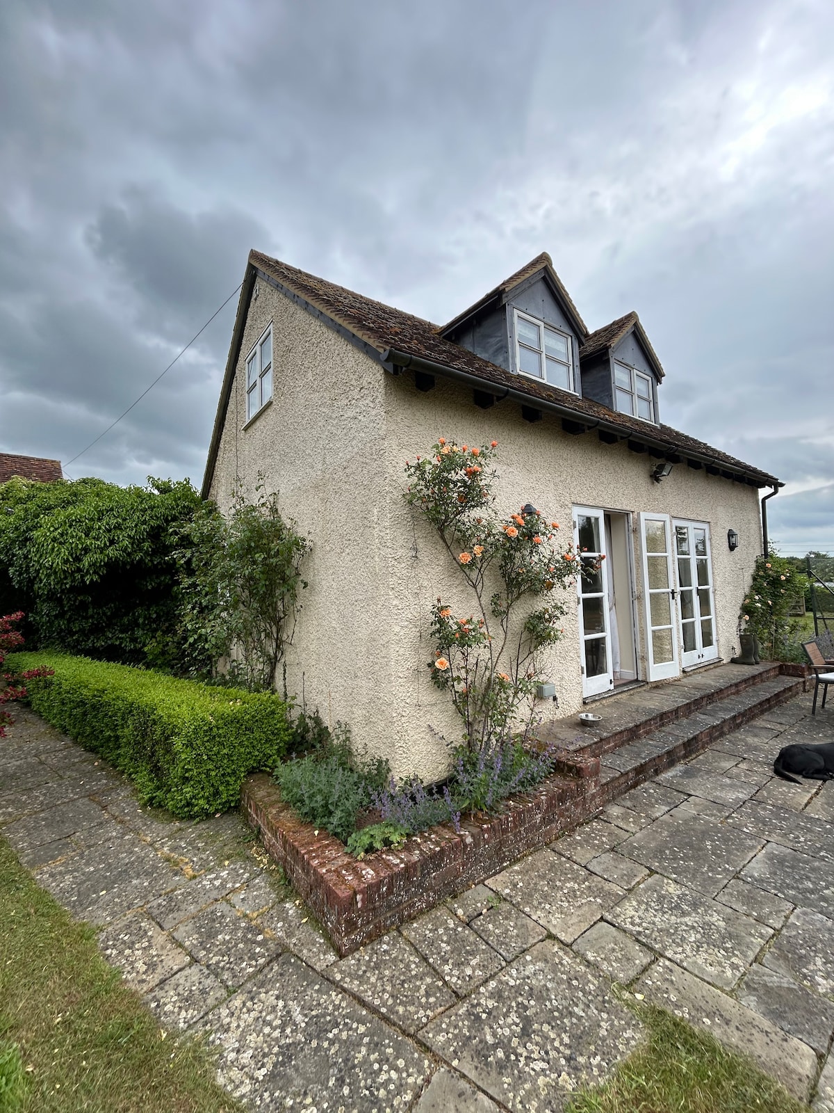 The cottage exterior is depicted, showcasing a charming building with a sloped roof and dual front windows. Flowering shrubs and neatly trimmed hedges frame the entrance, while a paved patio area leads to a set of double French doors that open to the interior.
