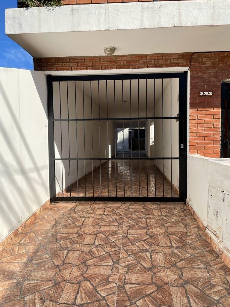 A gated entrance is shown, featuring a black metal gate that opens into a well-lit corridor. The floor is covered with patterned tiles, and natural light is visible at the far end, indicating a welcoming pathway to the interior.