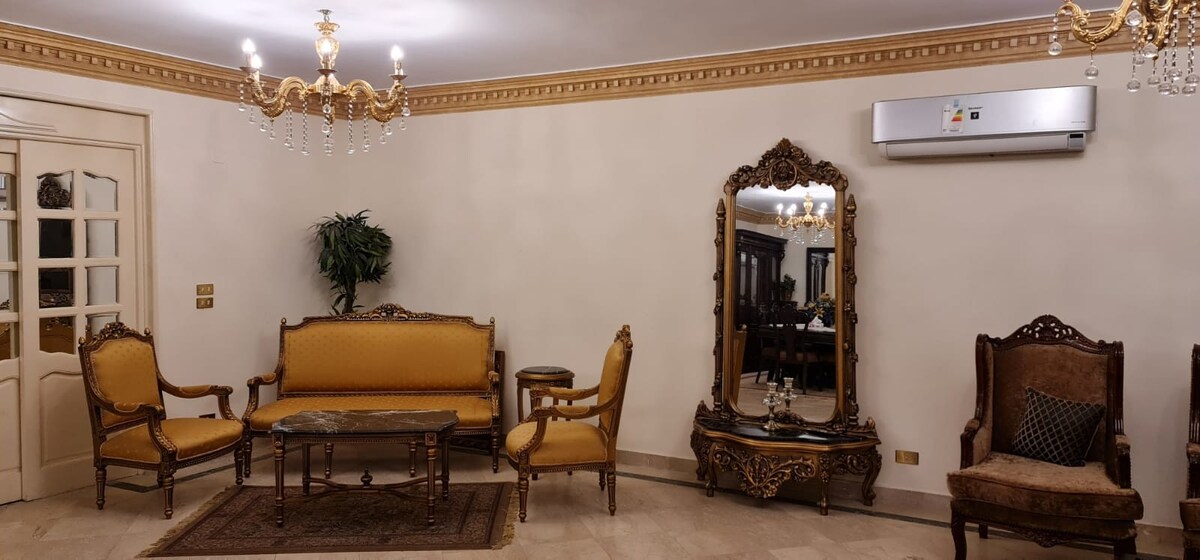 An elegantly furnished living space is presented, featuring ornately carved furniture in warm tones. A central wooden table is surrounded by two armchairs and a sofa, while a large mirror reflects the room's design. Decorative lighting hangs from the ceiling, enhancing the ambiance.