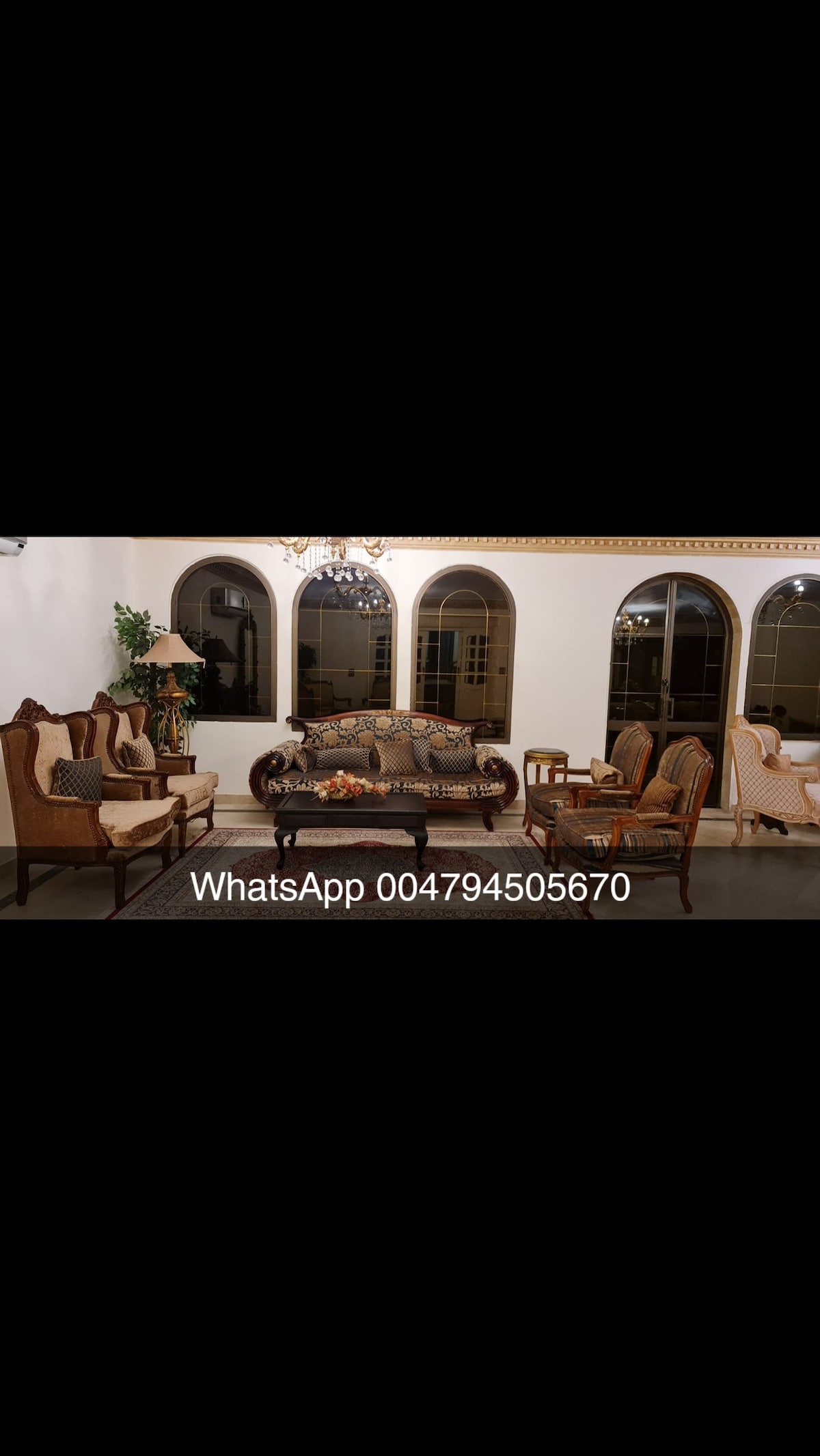 A spacious living area is arranged with multiple seating options, including a patterned sofa and several armchairs. Large arched windows allow natural light to fill the room, while decorative light fixtures enhance the space's elegance.
