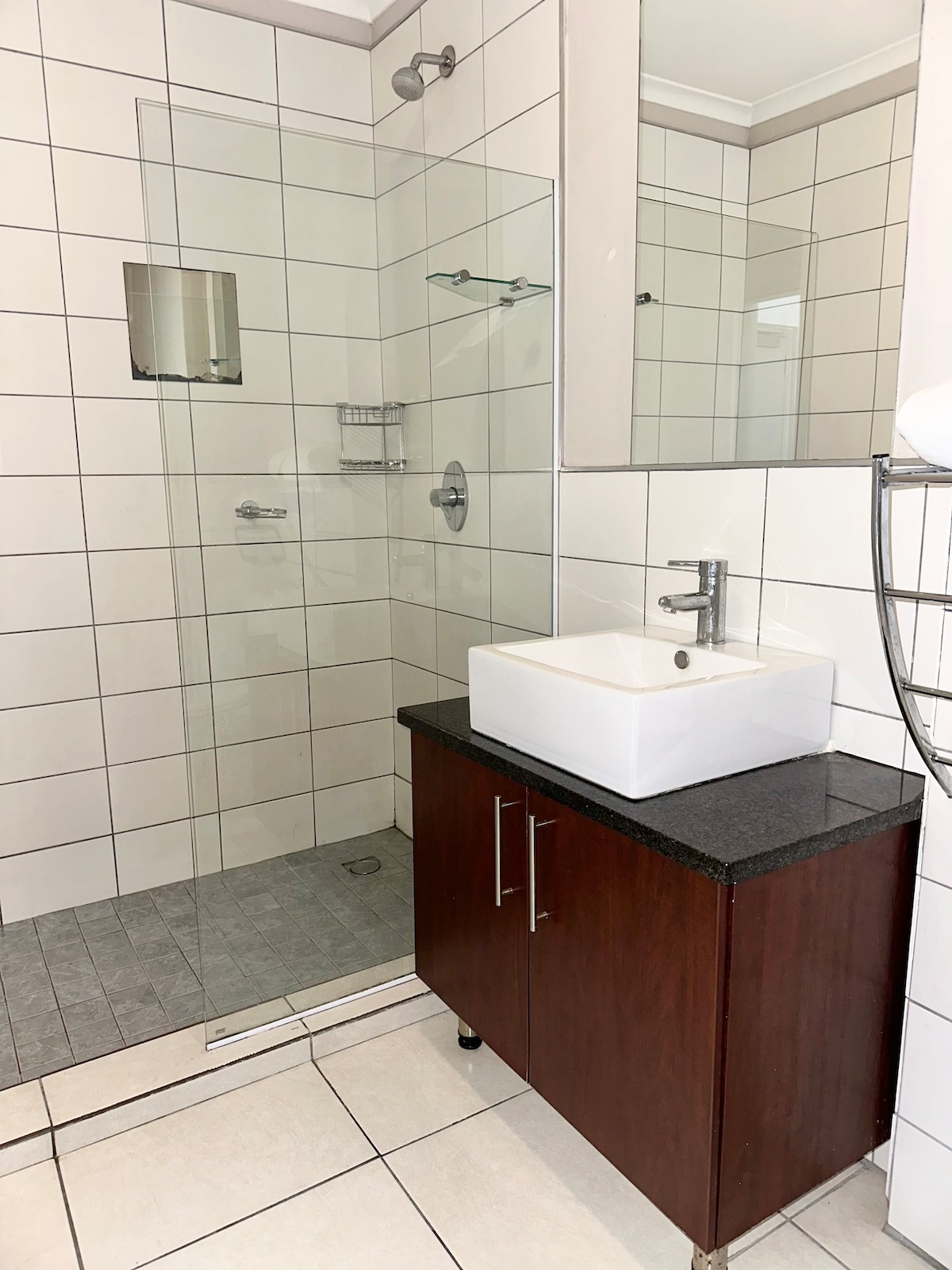 A modern bathroom features a walk-in shower enclosed by glass panels, complemented by a minimalist sink set on a dark countertop. White tiles create a clean and bright environment, while a wooden cabinet below provides practical storage.