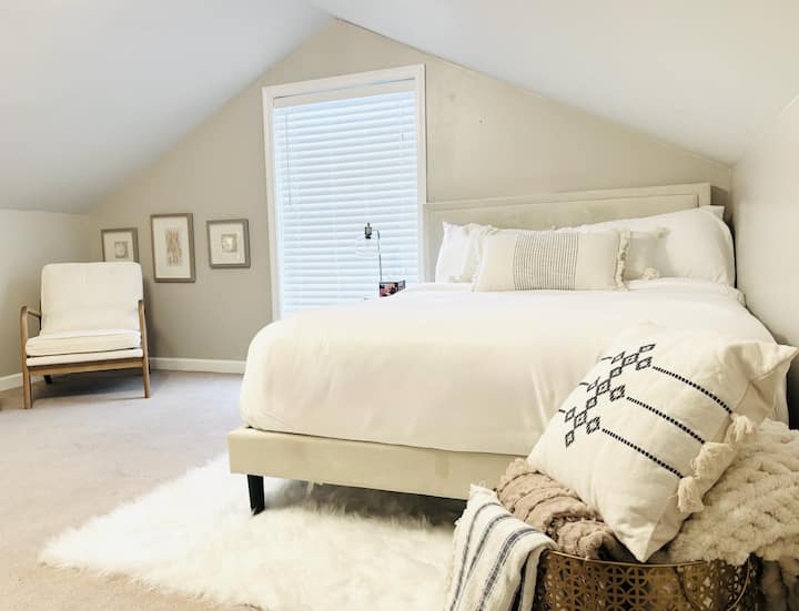 Hapësirë magjepsëse e stilit loft me tavane të pjerrët, shtroja të buta dhe dritë dielli që derdhet nga dritarja
