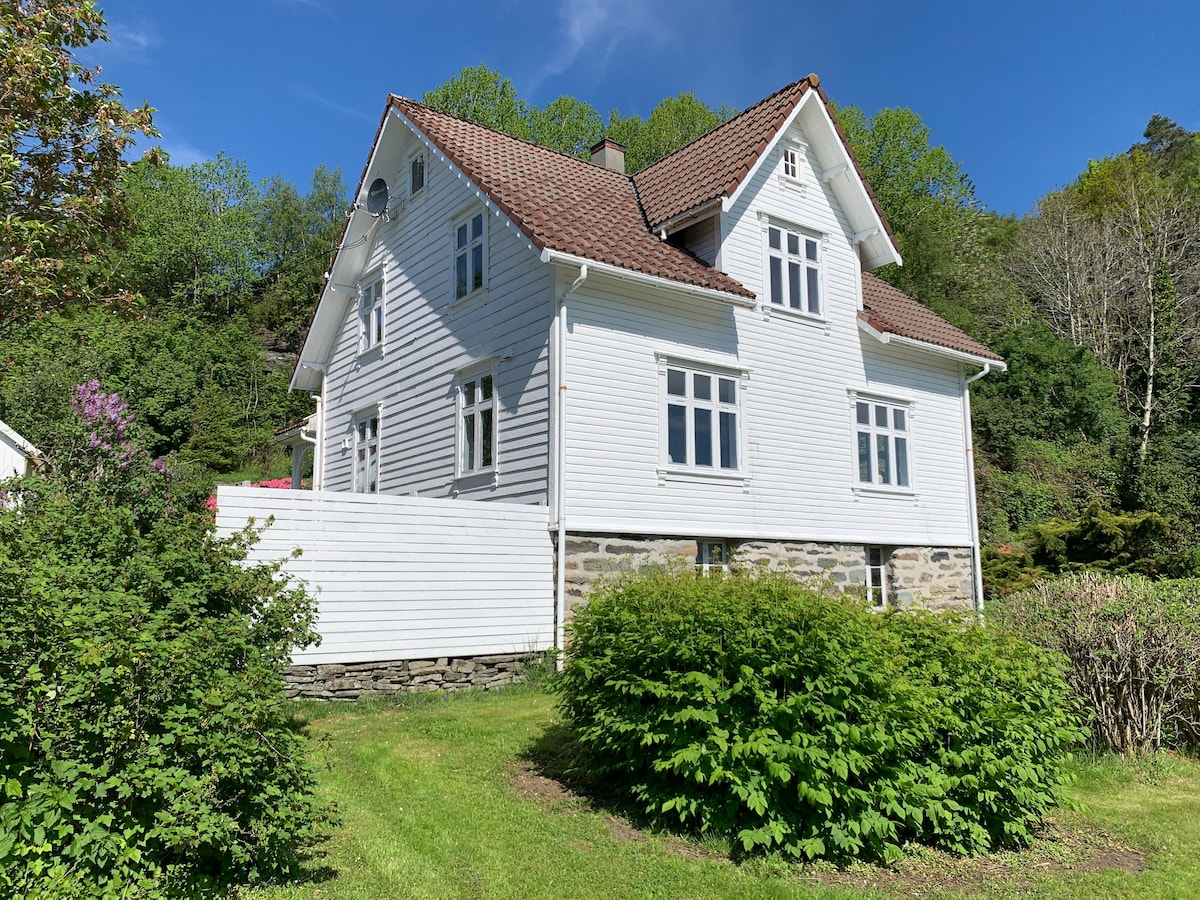 A two-story summer house is shown with a white exterior and a red-tiled roof. The structure is surrounded by lush greenery, including bushes and trees. Large windows reflect the sunlight, and a wooden fence partially encloses the property, enhancing its tranquil setting.