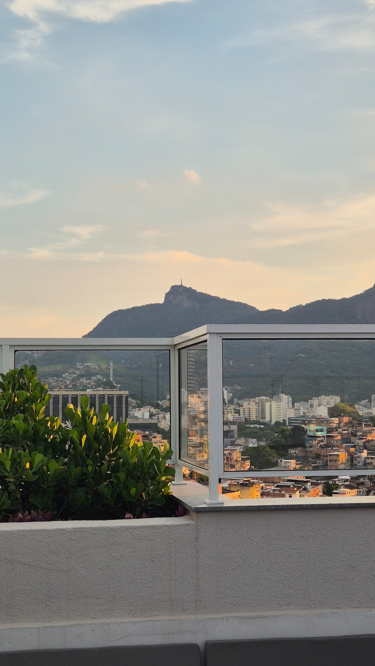 A rooftop view showcases the city skyline framed by a clear glass railing. Rolling mountains are visible in the background, with a prominent statue atop one peak. Lush greenery adds a natural touch to the space, enhancing the open atmosphere.