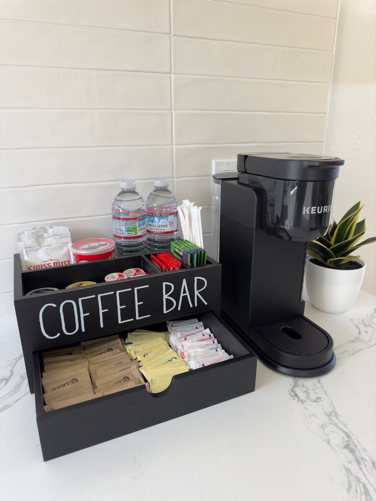 A coffee bar setup is displayed, featuring a coffee machine beside a wooden organizer. The organizer contains an assortment of coffee and tea supplies, including packets of sweeteners and napkins. Bottled water and drinking straws are arranged neatly on the countertop, with a small indoor plant adding a touch of greenery.