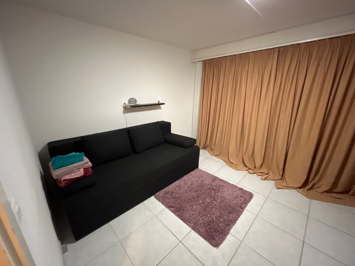 A compact room features a black sofa bed with neatly stacked towels beside it. A soft purple rug lies on the tiled floor, while light beige curtains cover a large window, providing a sense of warmth and privacy.