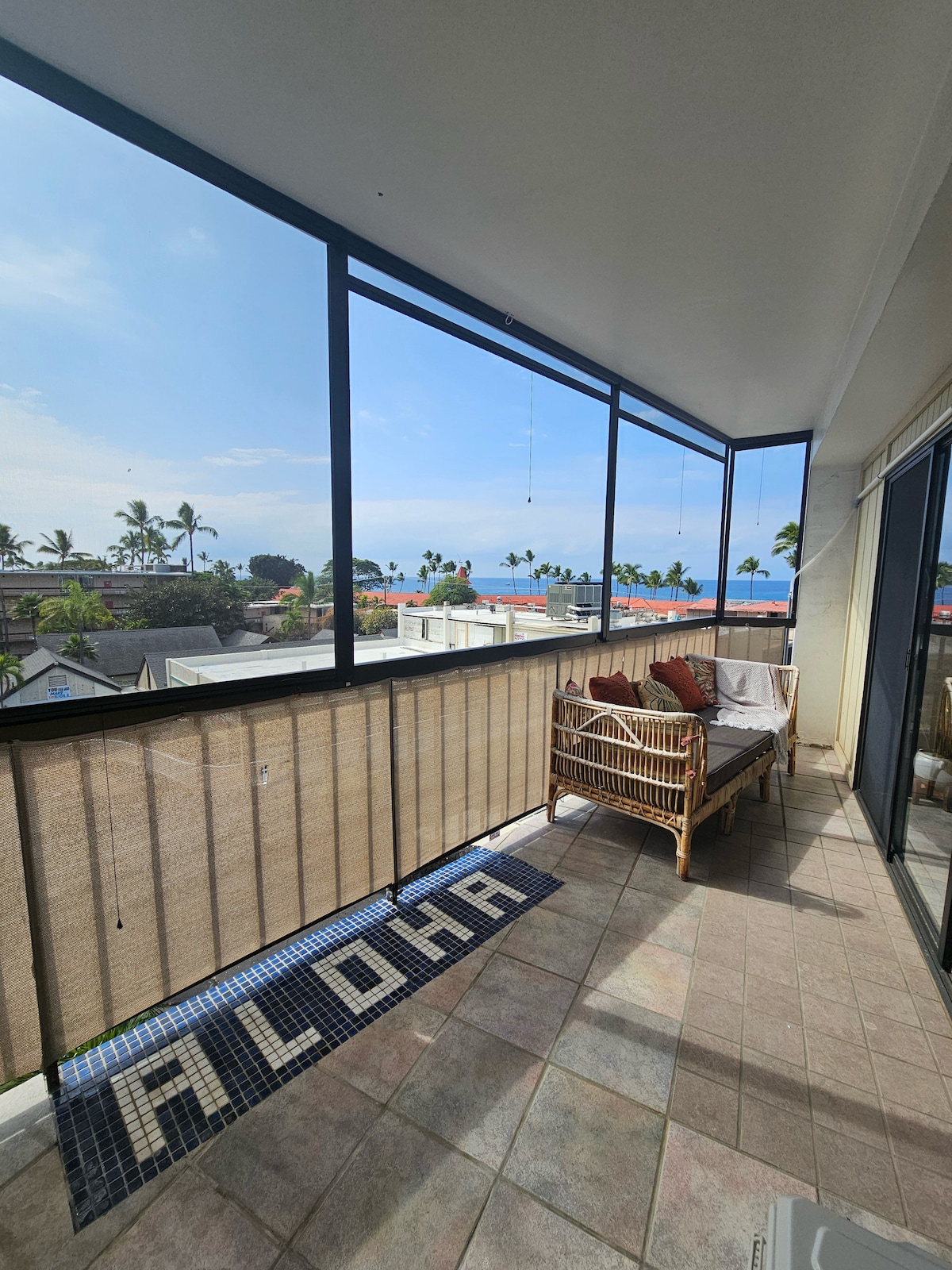 A private lanai is showcased with panoramic views of the ocean and surrounding palm trees. A comfortable seating area is present, featuring a cushioned bench. The tiled floor includes a welcome mat that reads 'Aloha,' enhancing the inviting outdoor space.