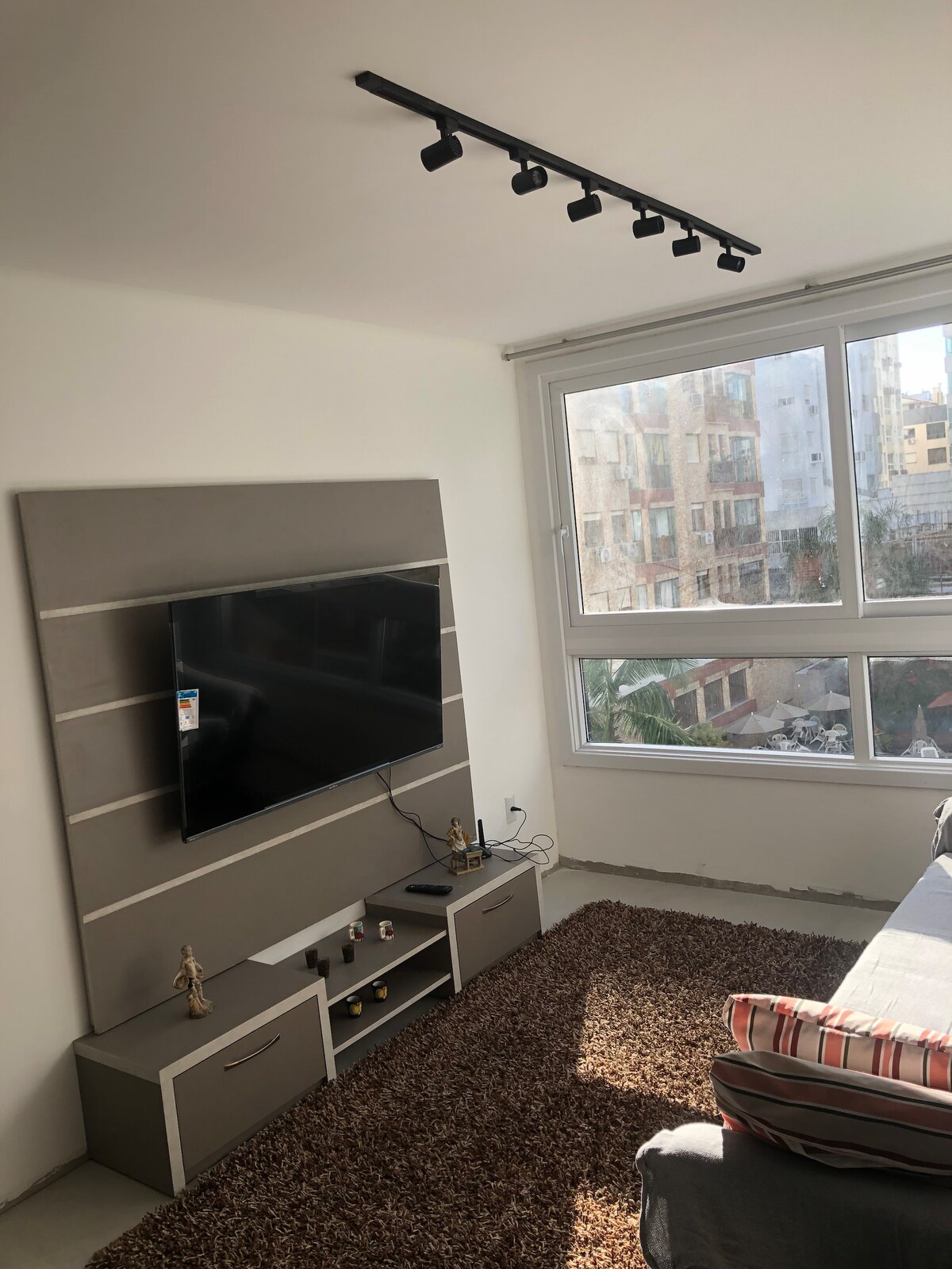 A well-lit living space is showcased, featuring a large flat-screen TV mounted on a stylish media unit. Natural light floods from the adjacent floor-to-ceiling windows, highlighting the plush area rug below. Soft furnishings provide comfort in the relaxed atmosphere.