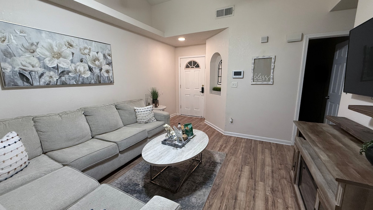 A modern living area features a spacious sectional sofa adorned with decorative pillows. A round coffee table sits centrally, holding a small decorative tray. Light-colored walls create an open feel, and a doorway leads to the entrance while a flat-screen TV is mounted on the wall.