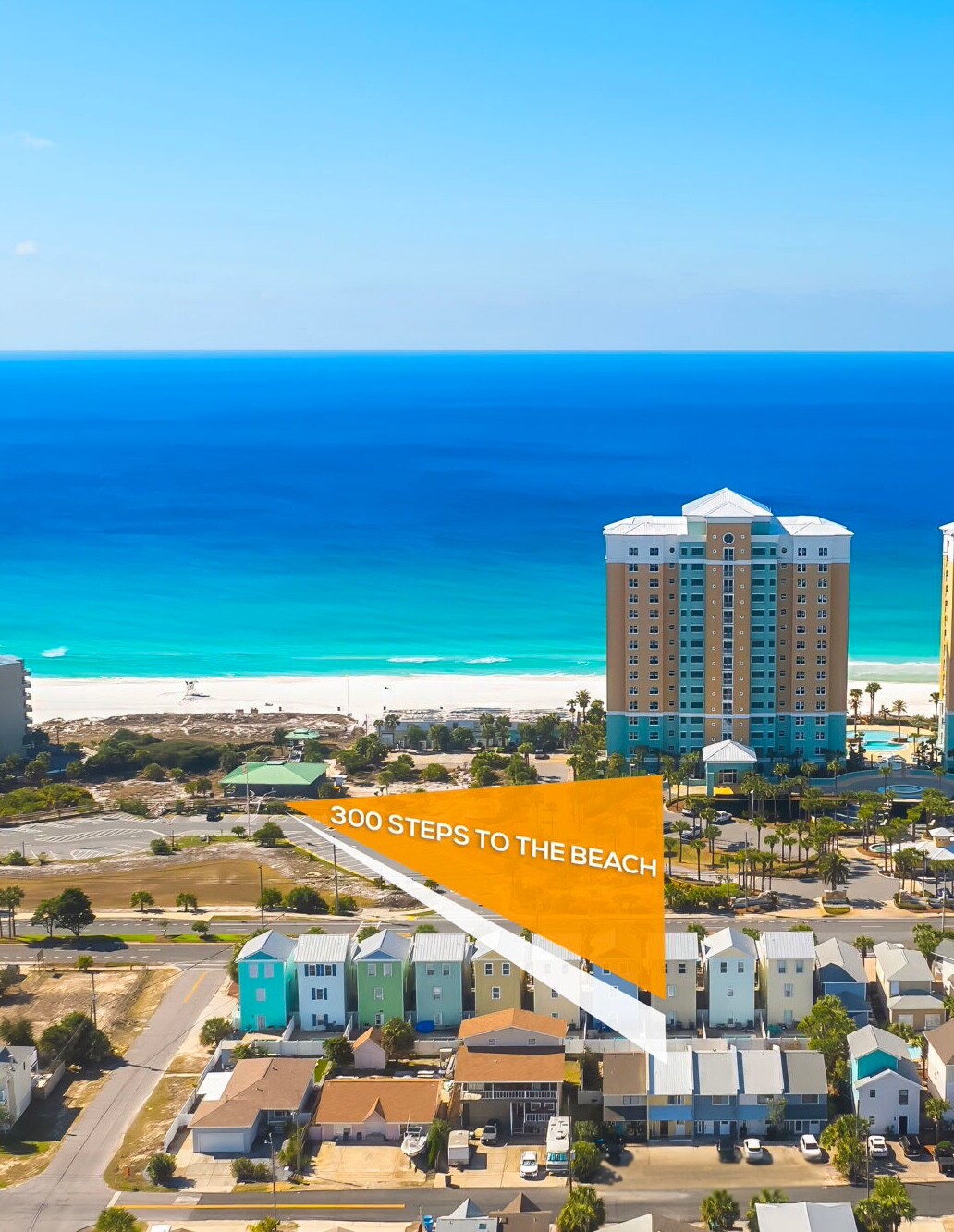 A scenic view showcases the coastline with turquoise waters meeting the sandy beach, just 300 steps from the location. Tall buildings stand in the background, while a mix of residential homes can be seen in the foreground, creating a picturesque coastal environment.