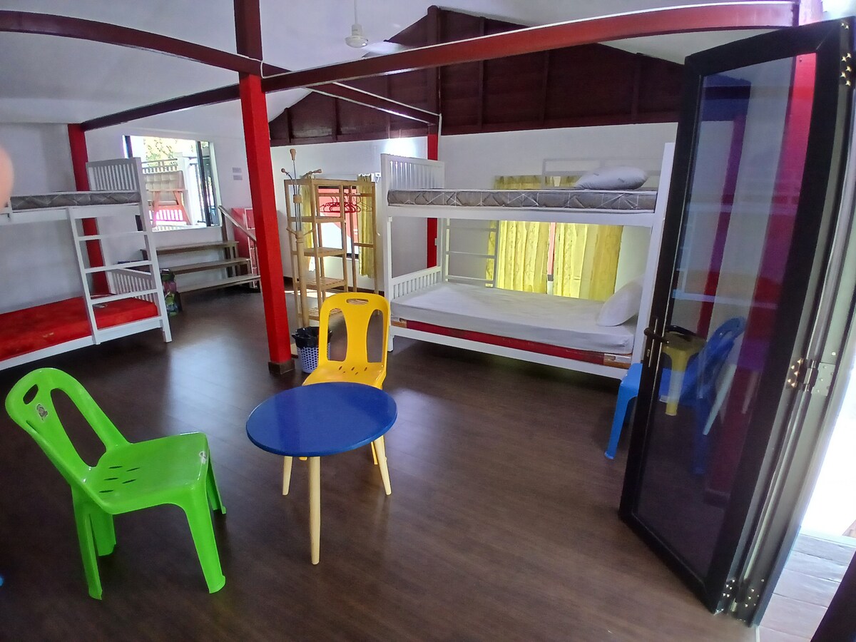 The interior features a bright, spacious room furnished with two bunk beds, accommodating four single beds, and a small blue table surrounded by plastic seating. Natural light streams in through large glass doors, highlighting the warm tones of the wooden floor.