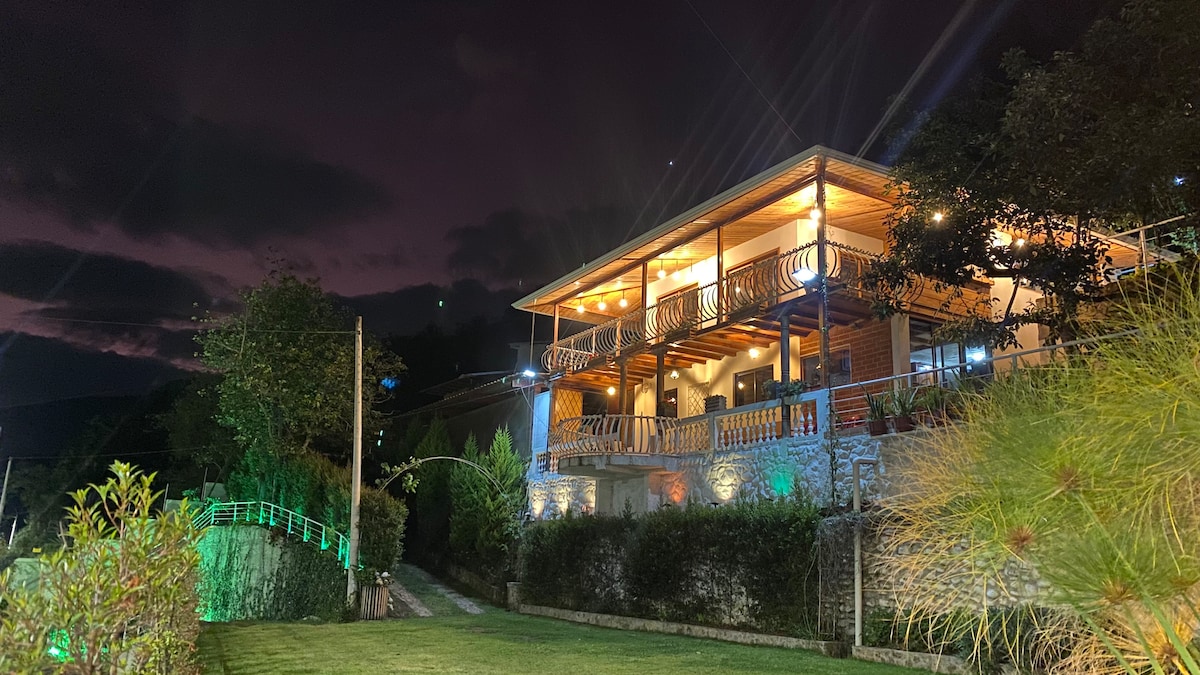The exterior of the house is illuminated at night, highlighting its wooden deck and stone features. A well-maintained garden with lush green plants surrounds the property, while soft lighting creates a welcoming ambiance. The sky above displays subtle hints of purple and stars.