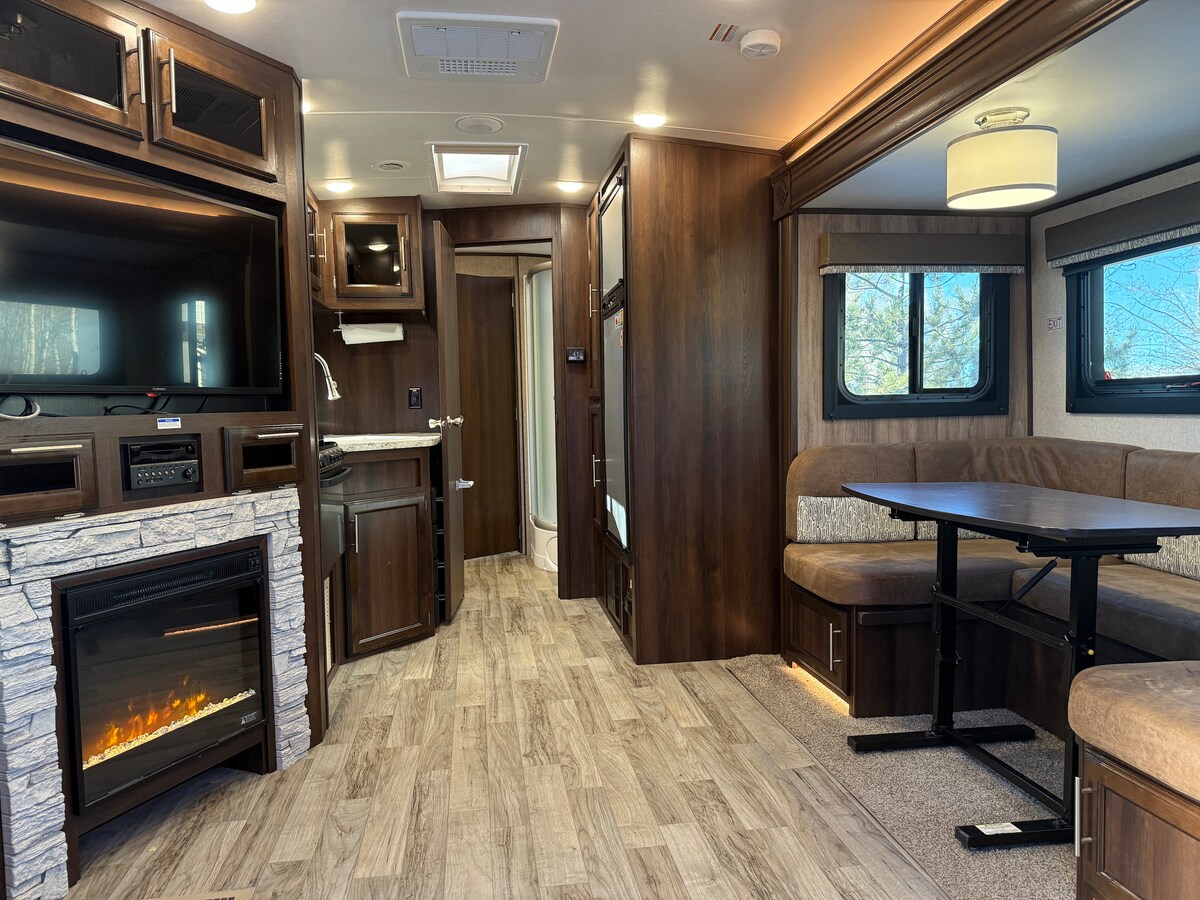 An indoor living space is displayed, featuring a dinette booth, equipped kitchen appliances, and a comfortable couch. A modern electric fireplace is positioned against the wall, and a flatscreen TV is mounted above a storage unit. Natural light filters through windows framed by warm-toned furnishings.