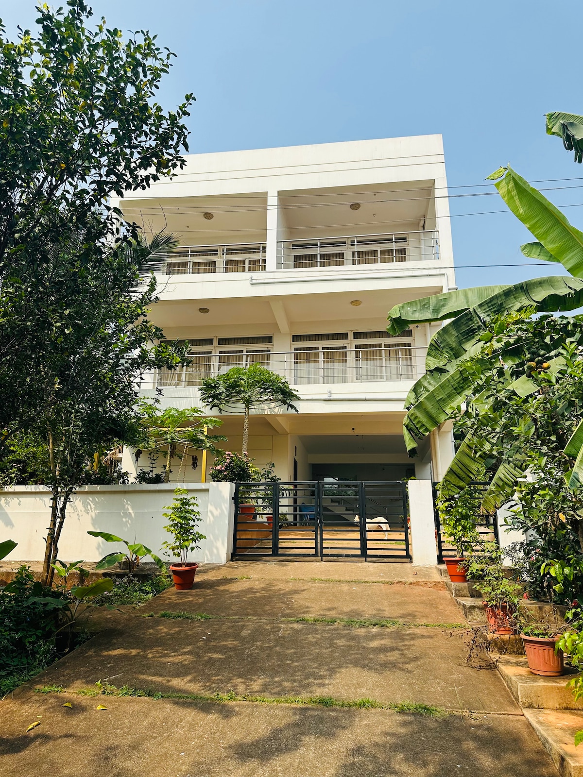 A modern three-story house is surrounded by lush greenery and potted plants at the entrance. The façade features multiple balconies with railing, providing a light and airy appearance. A driveway leads up to the main entry, inviting guests into the spacious retreat.