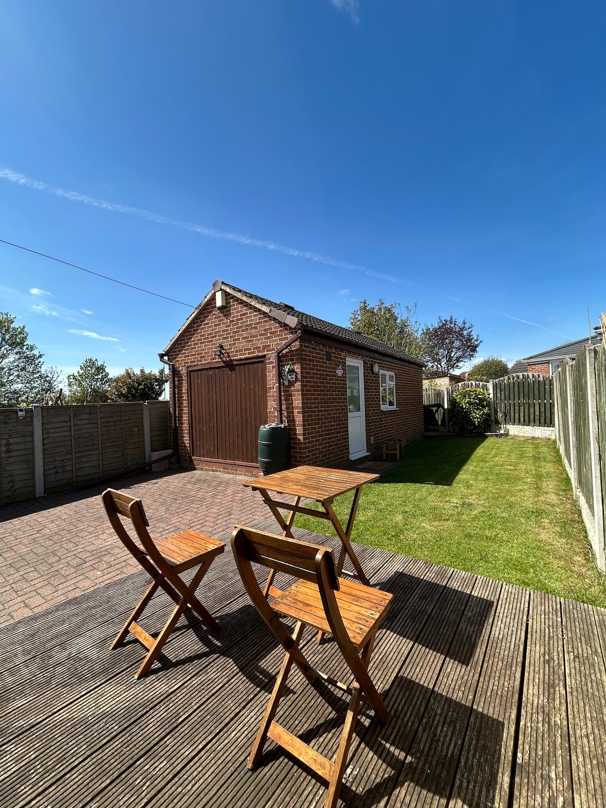 A private outdoor garden area is presented, featuring a patio deck with wooden furniture, including a table and two folding chairs. The well-maintained lawn and fenced boundaries create a serene environment, while a garden shed complements the outdoor space against a clear blue sky.