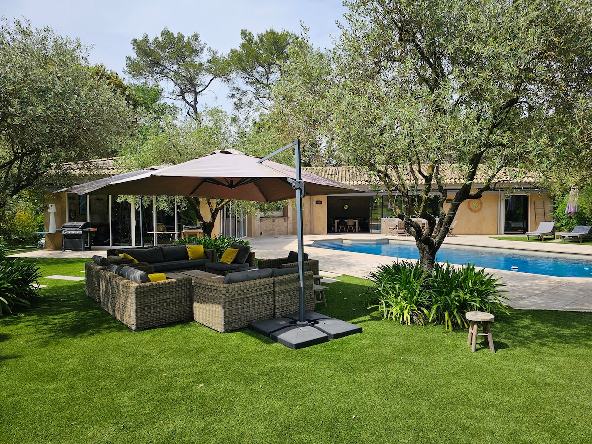 An inviting outdoor seating area is shown, featuring a large sectional sofa adorned with yellow cushions, situated under a large shaded umbrella. Lush greenery surrounds the space, with a sparkling pool in the background and a patio area visible in the distance.