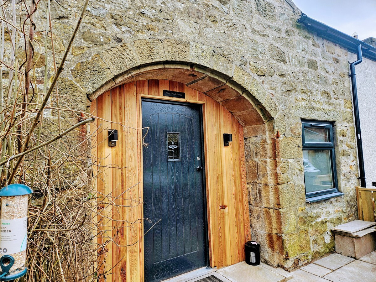 A rustic entrance to the tiny home is framed by a stone wall, showcasing an archway with a wooden door. The door features a sleek design and small wall lamps on either side, inviting guests into the space.