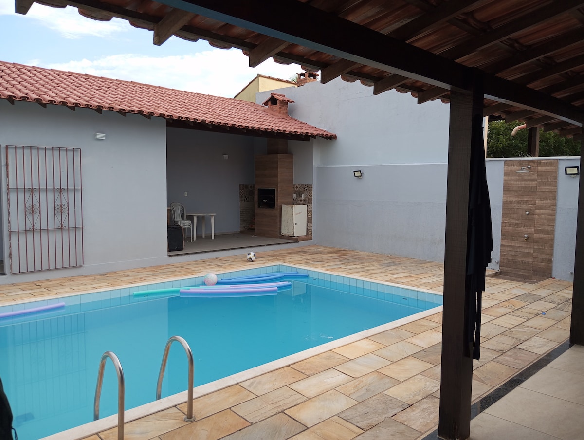 A private outdoor space is presented with a clear swimming pool, featuring colorful floats resting on the water's surface. A covered area alongside offers seating and a barbecue grill, while the tiled patio and stone wall contribute to the serene ambiance.