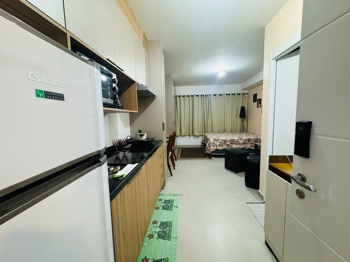 A modern studio kitchen is equipped with a refrigerator, induction stove, and various essential appliances on a black countertop. Beyond, a cozy sleeping area features a spacious bed with patterned bedding. Natural light enters through large windows, and curtains provide privacy.
