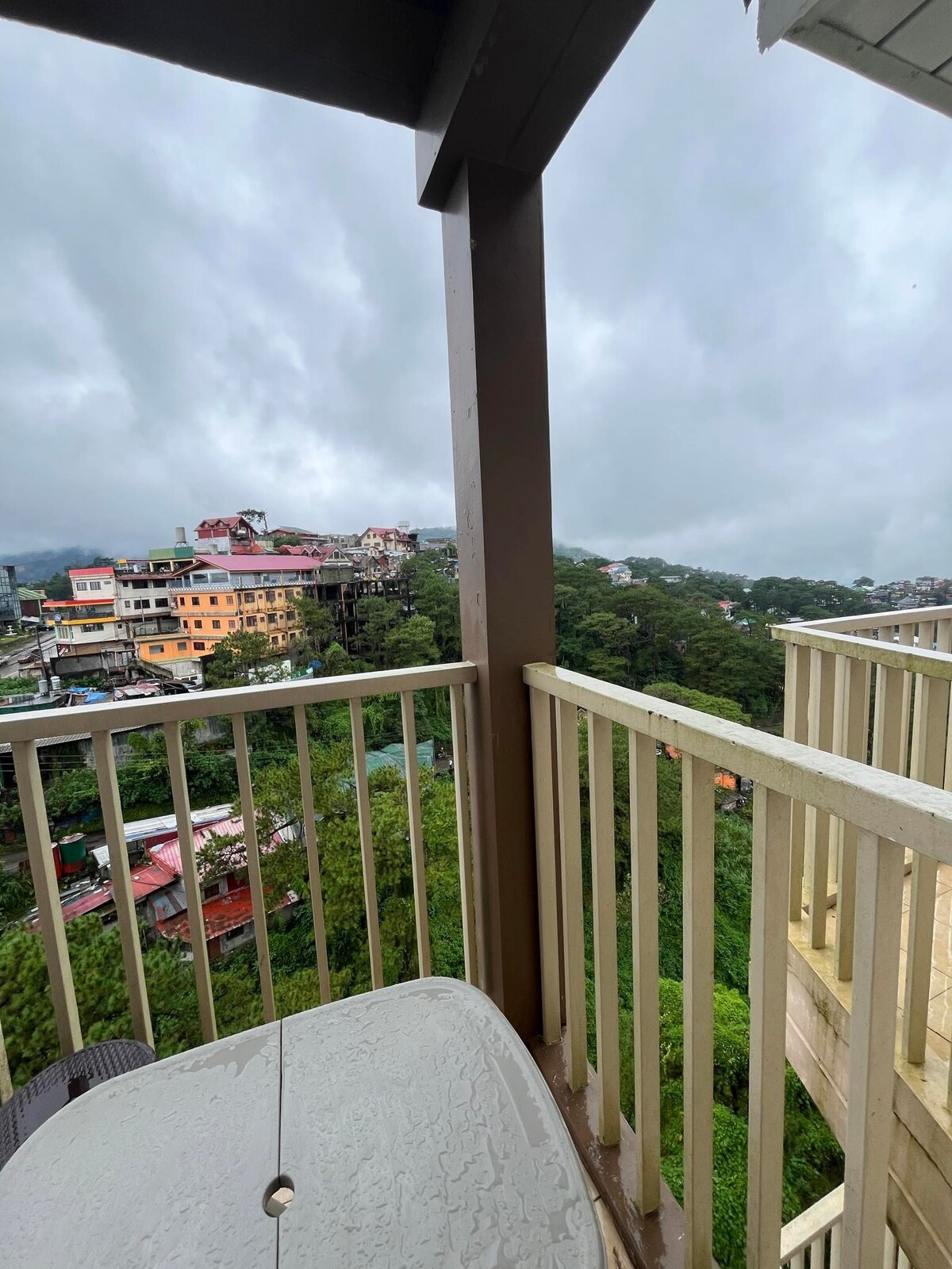 A balcony overlooks a lush hillside and a variety of colorful buildings below. The sky appears cloudy, contributing to a serene atmosphere. The railing is made of wooden slats, while a small table is positioned on the balcony, inviting moments of relaxation.