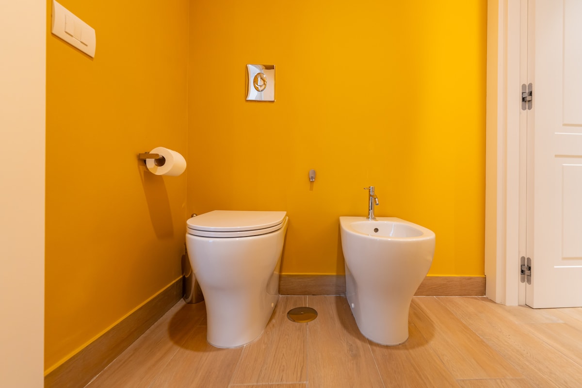 A contemporary bathroom features a bright yellow wall and modern fixtures. A toilet is situated next to a bidet, both set against a warm wooden floor. Soft lighting highlights the clean lines and minimalist design of the space.