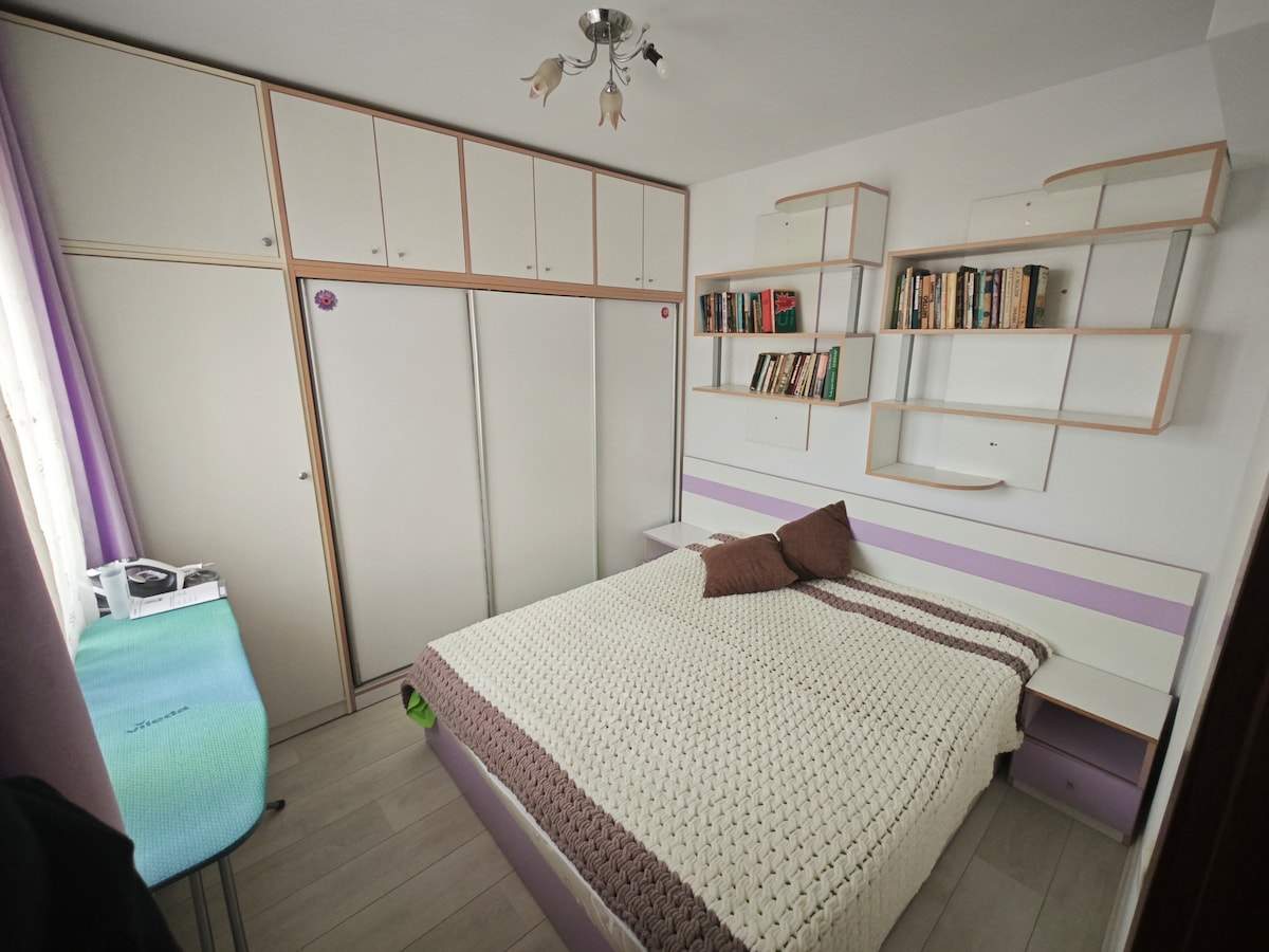 A well-organized bedroom is depicted, featuring a spacious bed with a textured blanket. Wall-mounted shelves hold books, while a small desk is positioned nearby. Natural light enters through a window adorned with sheer curtains, creating a bright and welcoming atmosphere.