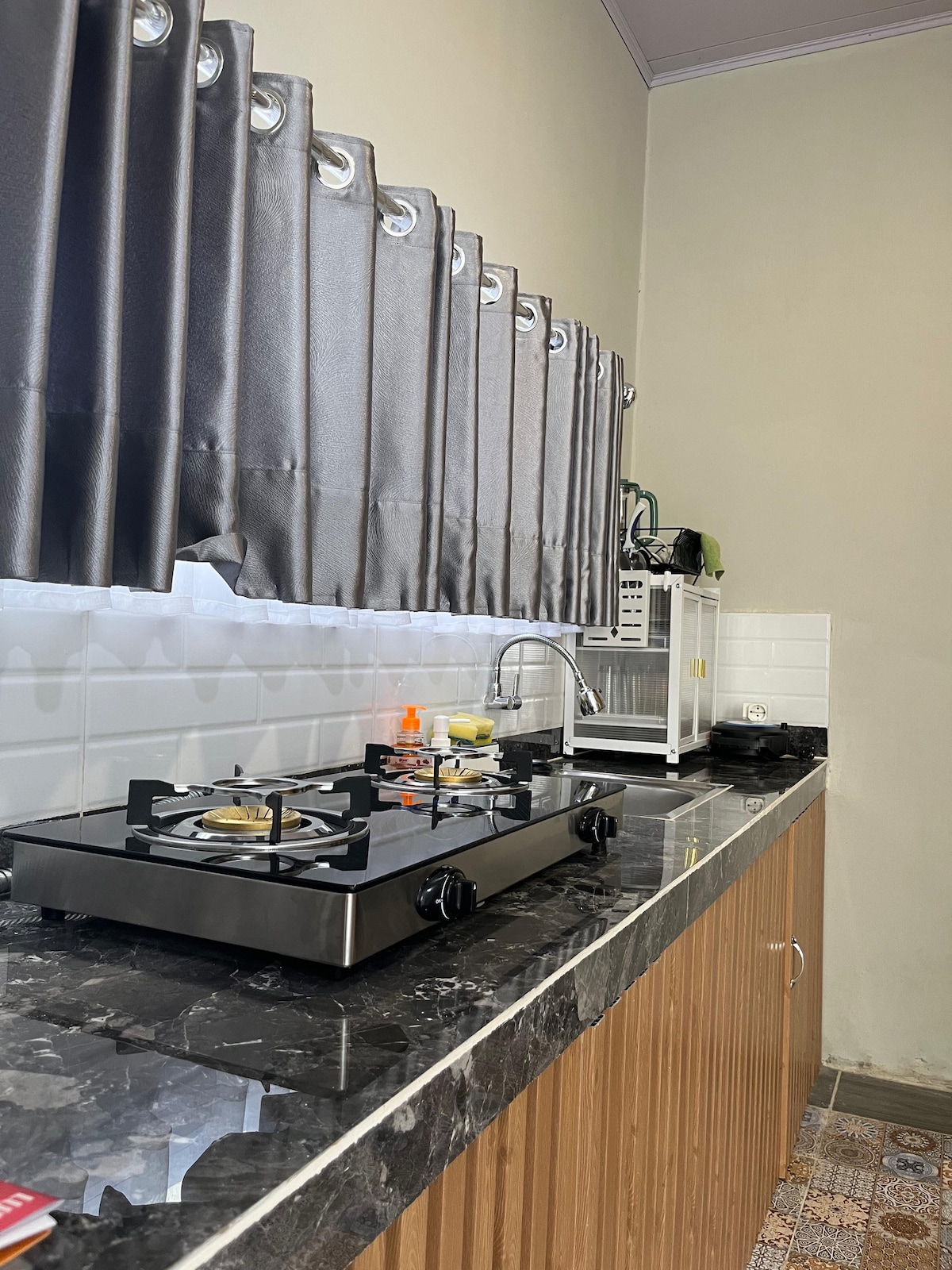 A modern kitchen is presented with sleek dark countertops and wooden cabinetry. A gas stove with multiple burners is visible, accompanied by a functional sink. Light gray curtains frame a window, allowing natural light to enhance the space.